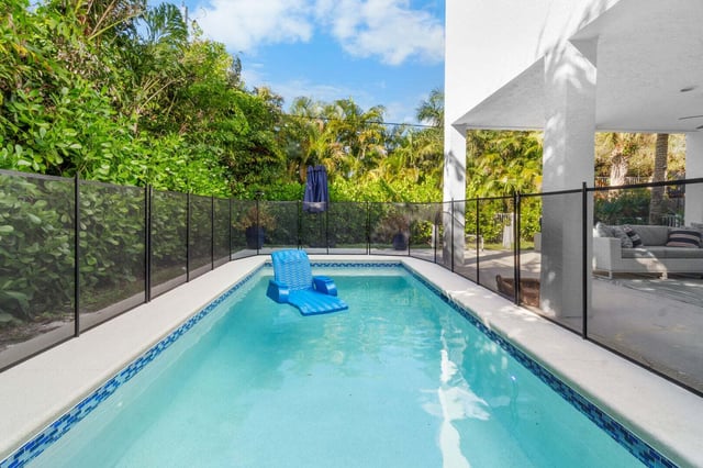 A backyard swimming pool with a blue lounger, safety fence and tropical palms under a bright sky