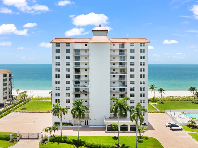 Aerial view of a white beachfront condo tower with orange roof, palm trees and turquoise Gulf water behind