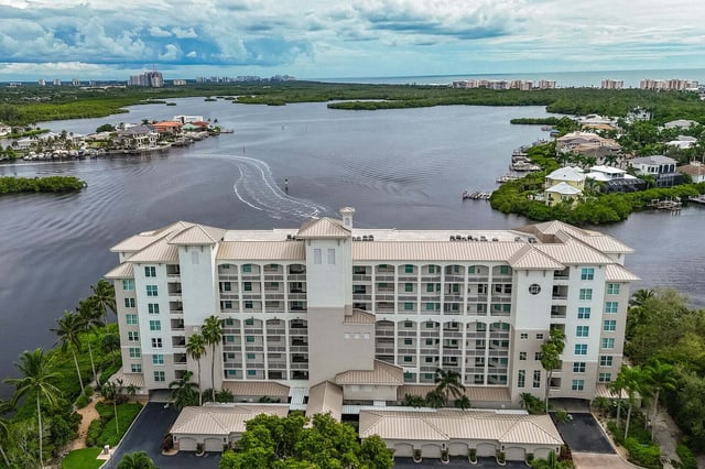 Aerial view of a waterfront condo building with metal roof overlooking a wide bay, boat wake and mangroves