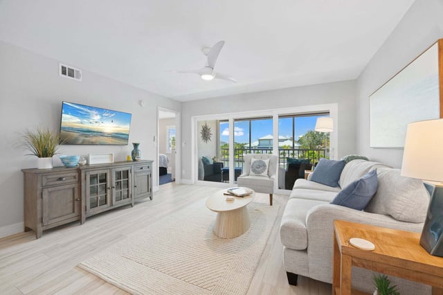 Bright coastal living room with white sofa, round coffee table, wall-mounted TV and sliding doors to a balcony