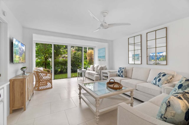 Bright coastal living room with white sectional, glass coffee table and sliding doors to a green garden