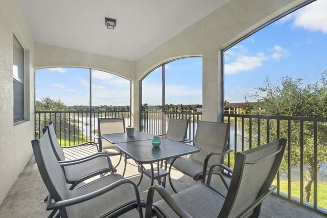 Screened balcony with patio table and four chairs overlooking a lake and palm trees under blue sky