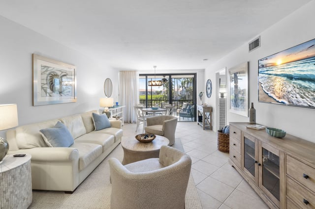 Bright coastal condo living room with cream sofa, blue pillows, tile floors and sliding doors to a palm-shaded lanai
