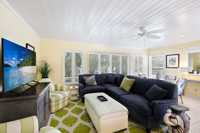 Bright coastal living room with navy sectional sofa, green patterned rug, plantation shutters and beadboard ceiling fan