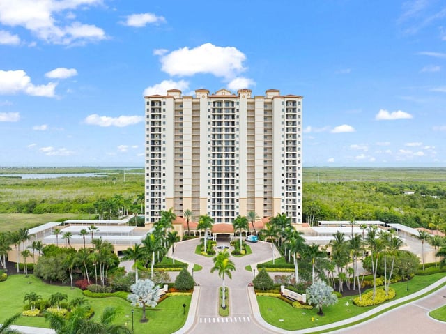 Aerial view of a high-rise condominium tower with palm-lined circular drive and mangroves under a blue sky