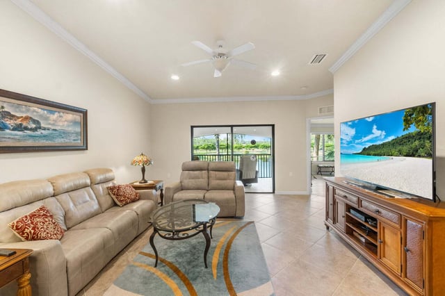 Bright living room with tan leather sofas, glass coffee table, large TV and sliding doors to a lake view lanai