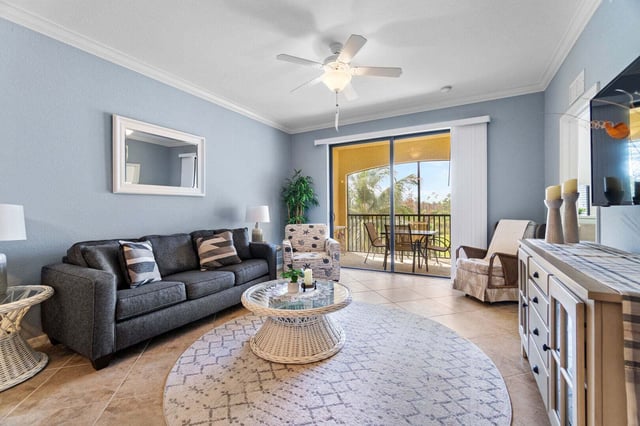 Bright condo living room with gray sofa, ceiling fan, round rug, and sliding doors to a sunny balcony