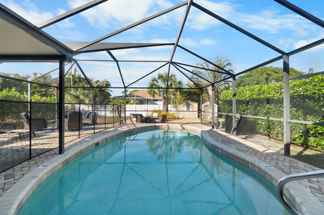 Screened lanai enclosing a turquoise pool with paver deck, patio chairs and palms under a blue sky