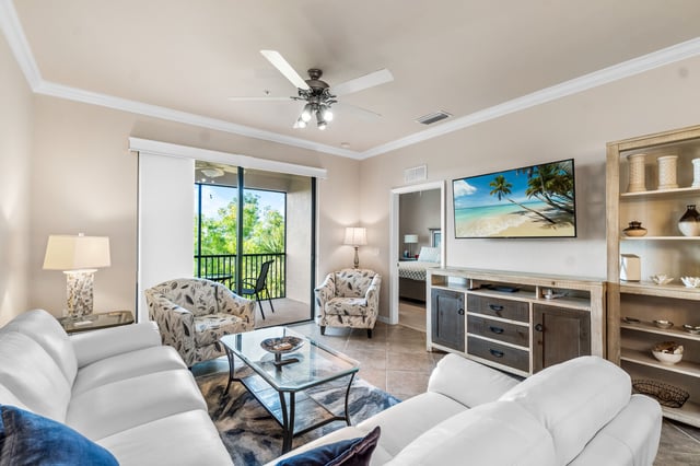 Bright condo living room with white sofas, ceiling fan, wall-mounted TV and sliding doors to a screened balcony