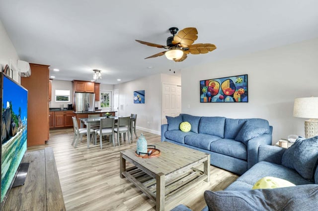 Open-plan living room with blue sofas, palm-leaf ceiling fan and wood coffee table beside a dining area and kitchen