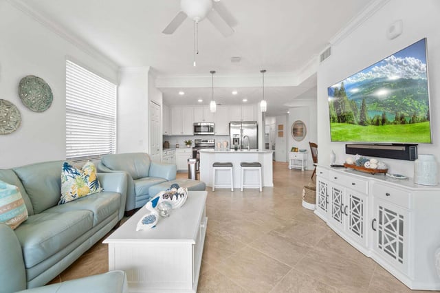 Open-concept living room with seafoam leather sofas, ceiling fan, wall-mounted TV and white kitchen island beyond