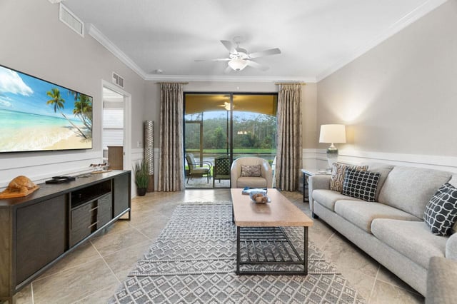 Condo living room with gray sofa, wall-mounted TV, ceiling fan and sliding doors to a screened lanai