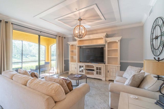 Bright living room with beige sofas, large TV in a built-in wall unit, and sliding doors to a screened lanai