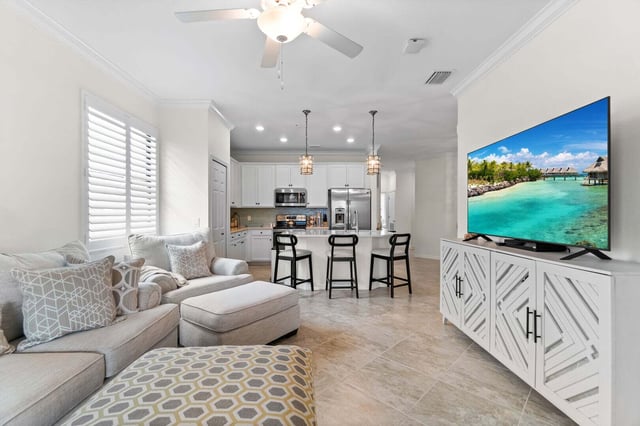 Bright open-concept living room with sectional sofa, wall-mounted TV and white kitchen with bar stools beyond