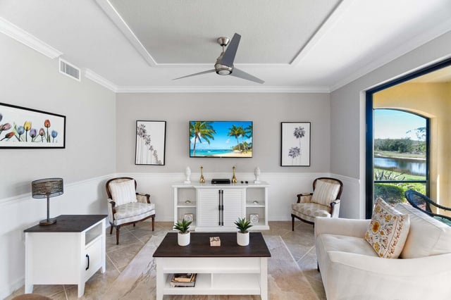Bright living room with ceiling fan, wall-mounted TV, white armchairs and sofa, and arched doorway to a lake view