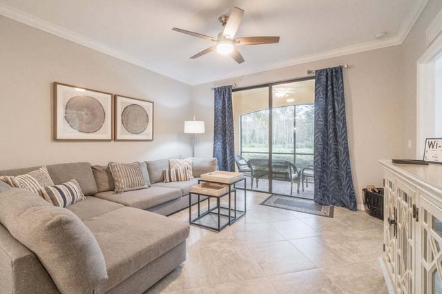 Bright condo living room with a gray sectional sofa, ceiling fan, and sliding doors to a screened lanai overlooking greenery