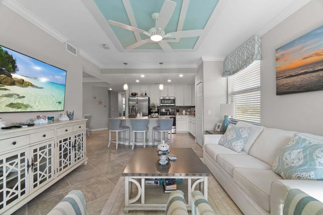 Coastal-style living room with white sofa, turquoise tray ceiling, ceiling fan and kitchen bar stools beyond