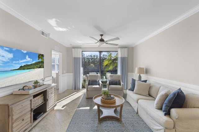 Bright condo living room with white sectional sofa, ceiling fan, wall-mounted TV and sliding doors to a palm-shaded patio