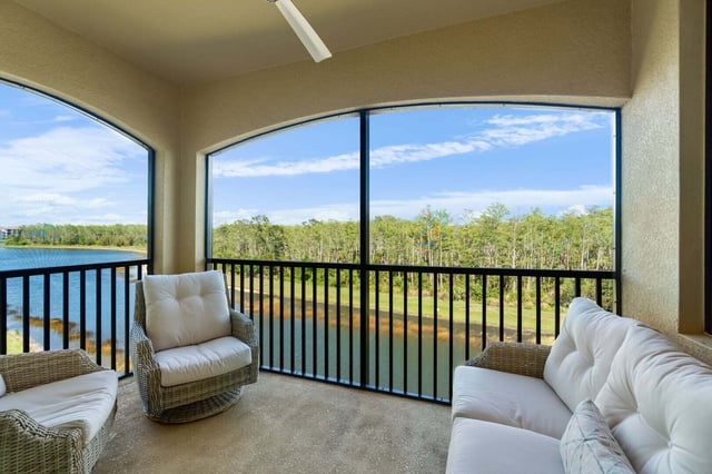 Screened lanai with white wicker chairs and sofa overlooking a lake and preserve treeline under blue sky