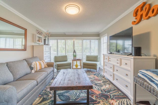 Bright vacation rental living room with gray sofa, tropical print rug, wood coffee table, and wall-mounted TV
```