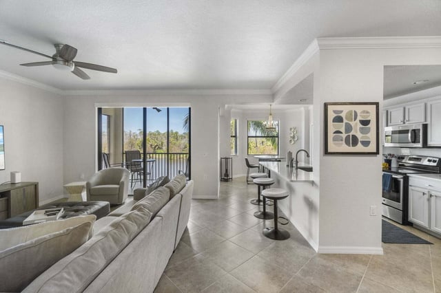 Open-plan condo living room with sectional sofa, ceiling fan, breakfast bar stools and a balcony overlooking greenery
