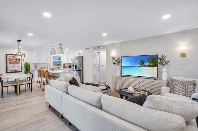 Bright open-plan living room with cream sectional, wall-mounted TV and white kitchen with barstools beyond