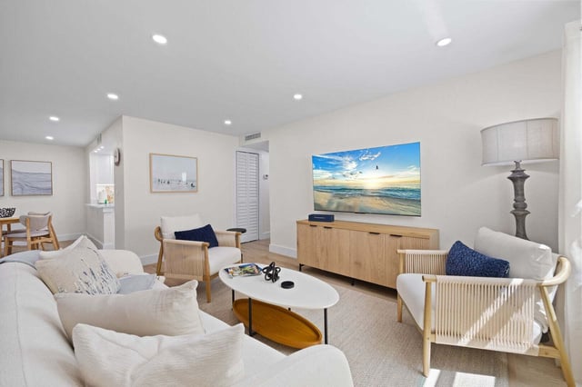 Bright coastal living room with white sofa, woven armchairs, oval coffee table and wall-mounted TV showing a beach scene