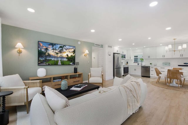 Open-plan living room with white sofas, wall-mounted TV and sage walls flowing into a bright white kitchen