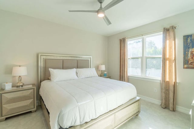 Bright bedroom with a king bed in white linens, upholstered headboard, ceiling fan and sunlit curtained window