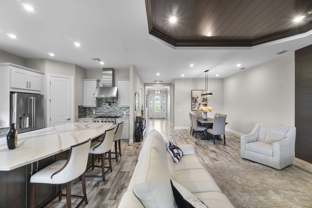 Open-concept living space with white marble kitchen island, bar stools, dining table and a dark wood tray ceiling