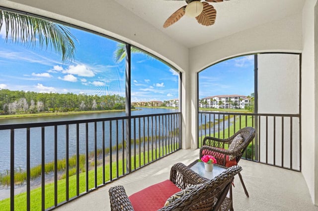 Screened lanai with wicker chairs and ceiling fan overlooking a lake, palm trees and condo buildings