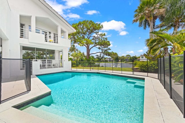 Turquoise backyard pool with safety fencing beside a white two-story waterfront home lined with palms
and blue sky