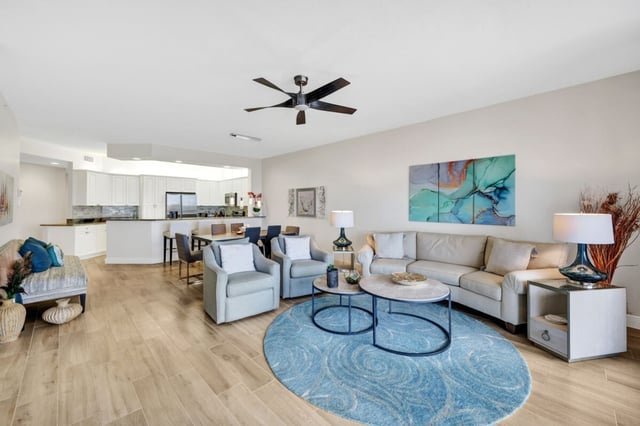 Open-plan condo living room with beige sectional, blue round rug, ceiling fan and white kitchen beyond