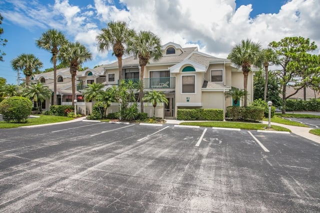Two-story condo building with balconies and palm trees beside an empty parking lot under a blue sky
<br>
