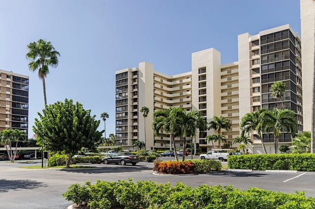 High-rise beige condo towers framed by palms and trimmed hedges above a parking lot under clear blue sky