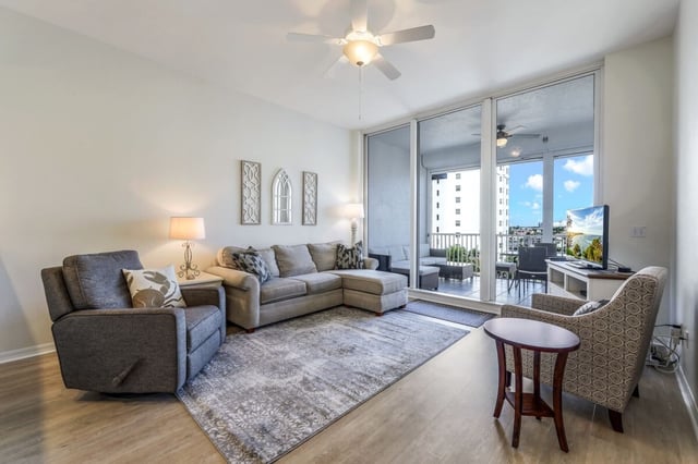 Condo living room with gray sectional sofa, ceiling fan and sliding glass doors opening to a sunny balcony