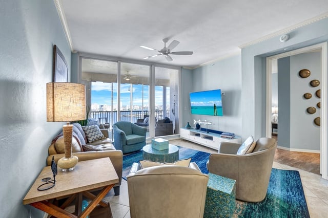 Coastal-style condo living room with blue rug, wall-mounted TV, ceiling fan, and balcony doors framing a water view