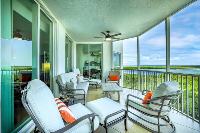 Screened condo balcony with cushioned lounge chairs, orange pillows and a ceiling fan overlooking mangroves and water
```