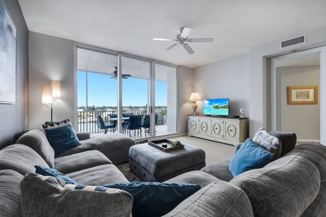 Condo living room with a large gray sectional, ottoman, ceiling fan and sliding doors to a balcony with water views