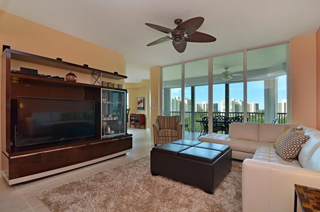 Condo living room with large flat-screen TV, sectional sofa, leather ottoman and floor-to-ceiling windows onto a balcony
