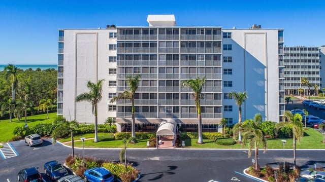 Aerial view of a white beachfront condominium tower framed by palm trees, parking lot and Gulf water beyond