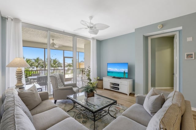 Bright condo living room with sectional sofa, palm-leaf ceiling fan, flat-screen TV and sliding doors to a screened balcony