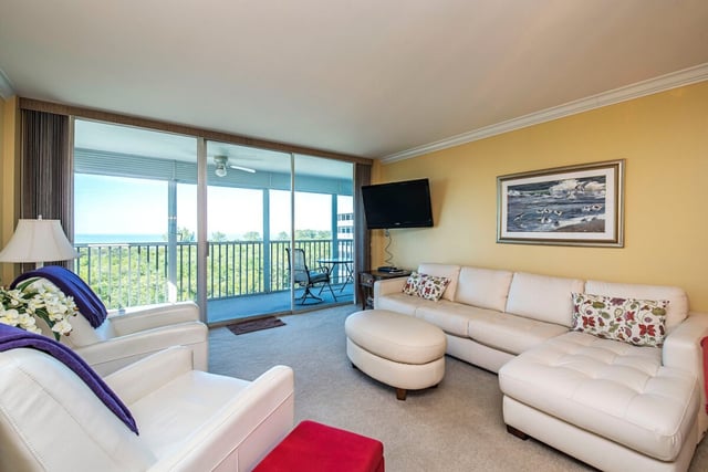 Condo living room with white sectional sofa, round ottoman and sliding doors opening to a screened balcony with treetop views