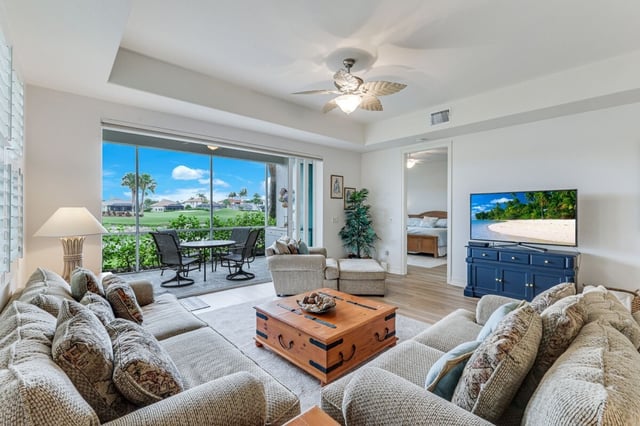 Bright condo living room with sectional sofa, wood trunk coffee table and sliding doors to a screened lanai overlooking a golf course