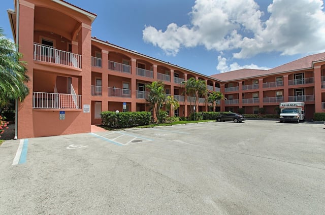 Three-story salmon-pink condo building with balconies, palm trees and an empty parking lot under blue sky