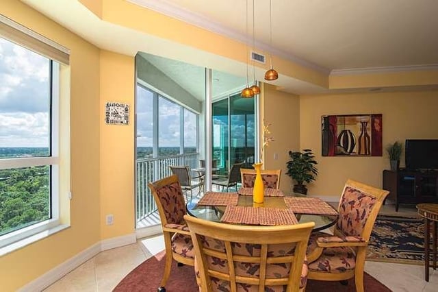 Sunlit high-rise condo dining area with glass table, cushioned chairs and floor-to-ceiling windows overlooking treetops