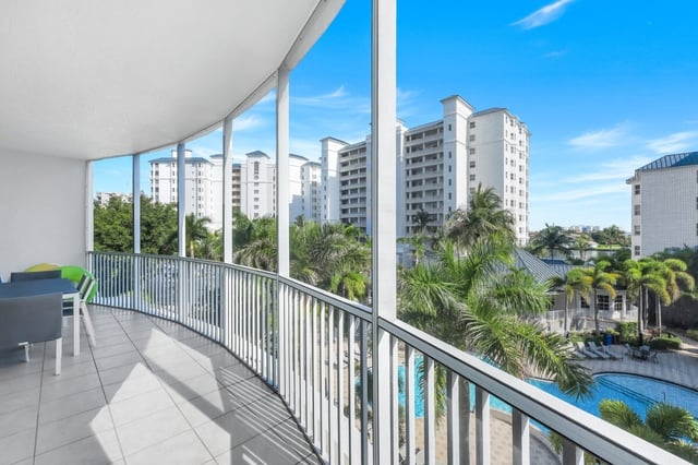 Curved condo balcony with white railing overlooking palm trees, a resort pool and high-rise towers under blue sky