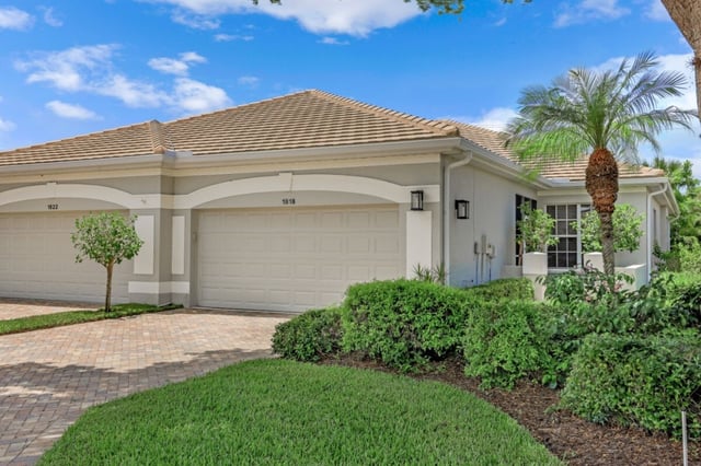 Single-story villa with tile roof, two-car garage, paver driveway and palm trees under blue sky