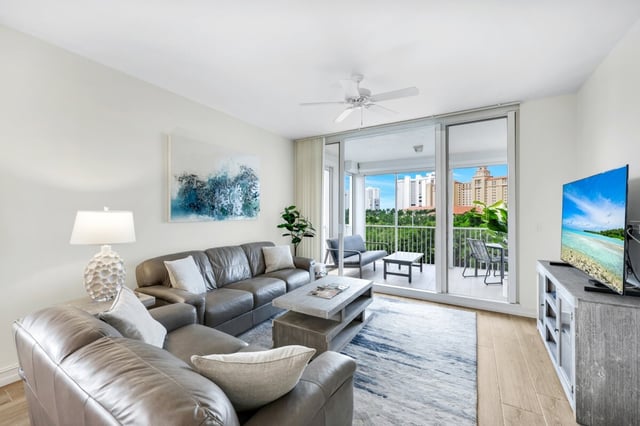 Bright condo living room with gray leather sofas, ceiling fan and sliding glass doors to a balcony with treetop views