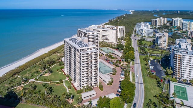 Aerial view of beachfront high-rise condo towers lining the Gulf shoreline with tennis courts and coastal road below